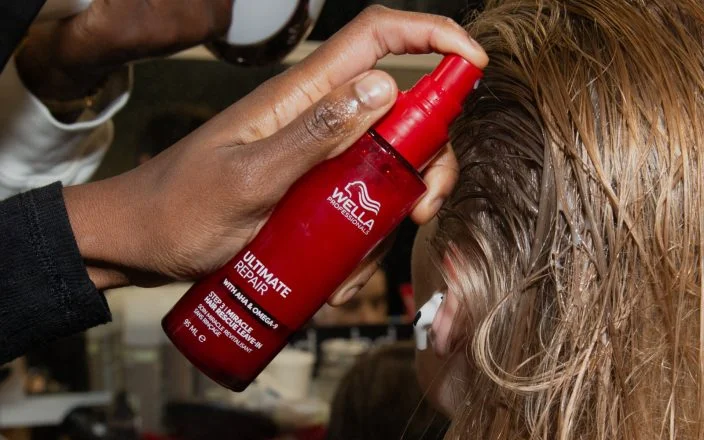 Hand holds a Wella spray bottle as it's squirted on light brown hair.