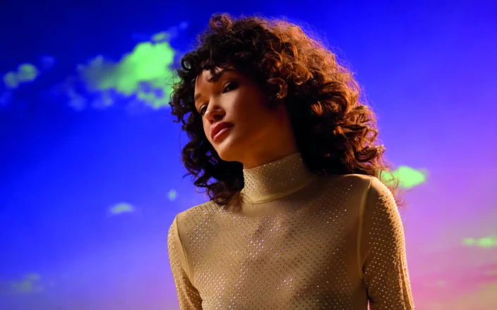 Woman with voluminous curly hair wearing a shimmering high-neck gown, seated on sand against a vivid sunset backdrop.