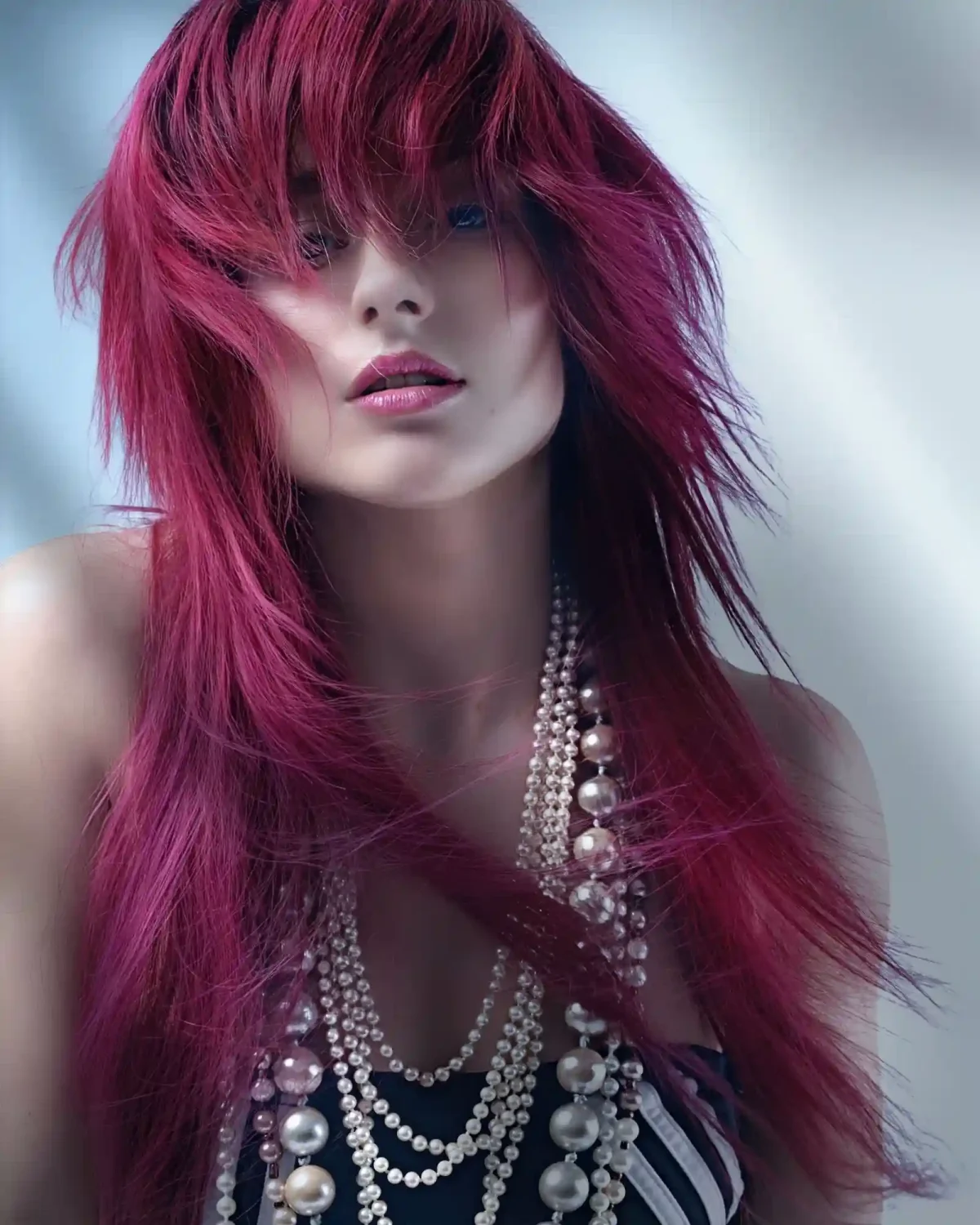 Editorial beauty portrait featuring vibrant magenta layered hair with textured fringe and pearl statement accessories.