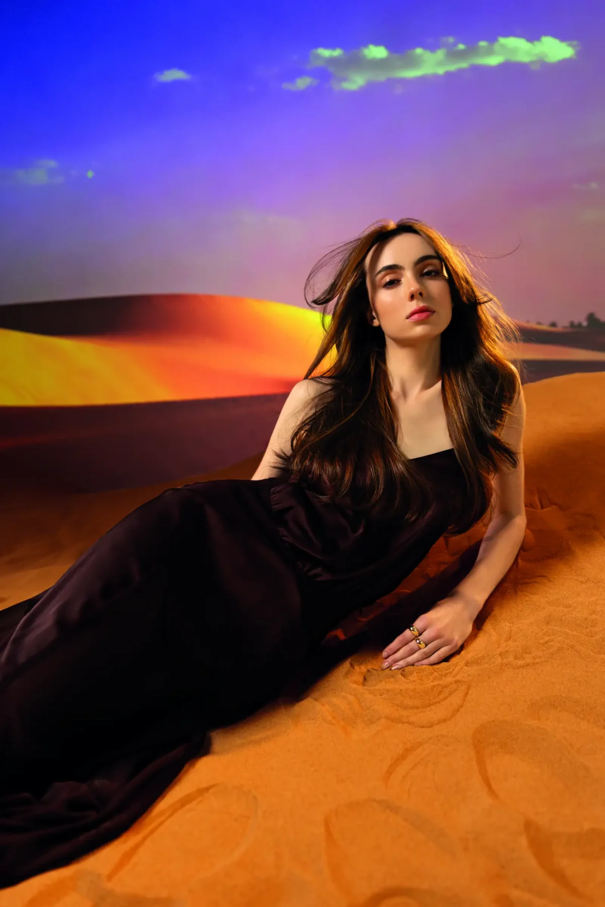 Woman with long glossy brunette hair reclining on desert sand in a black gown against a vivid sunset backdrop.