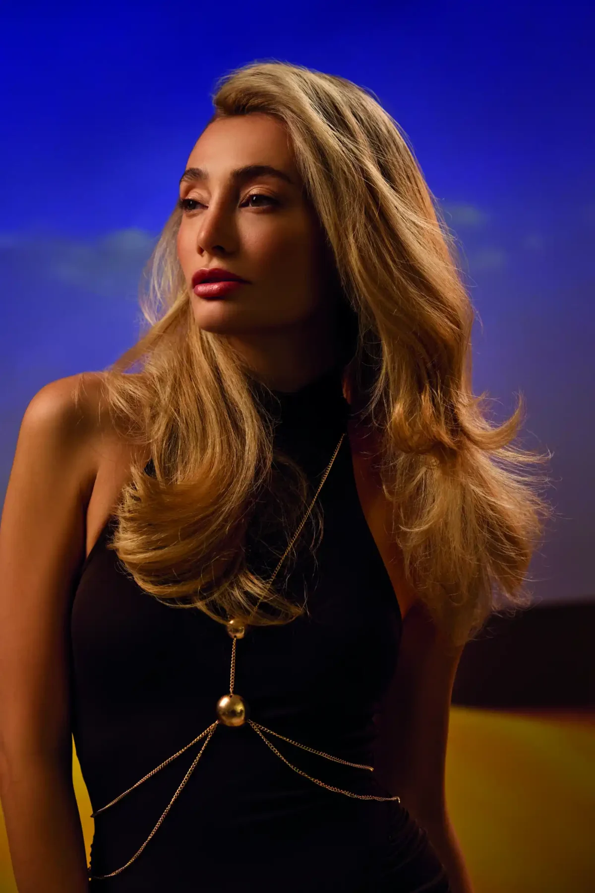 Woman with glossy honey blonde waves wearing a black dress against a dramatic sunset-inspired backdrop.