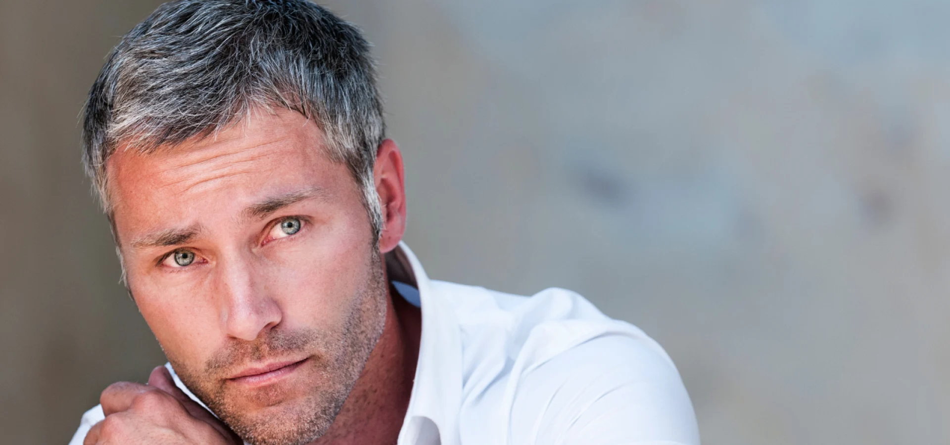 Close-up portrait of a man with short grey hair wearing a white shirt and wristwatch against a soft neutral background.