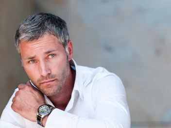 Close-up portrait of a man with short grey hair wearing a white shirt and wristwatch against a soft neutral background.