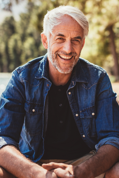 Smiling mature man with silver hair and beard wearing a denim shirt outdoors in natural sunlight.