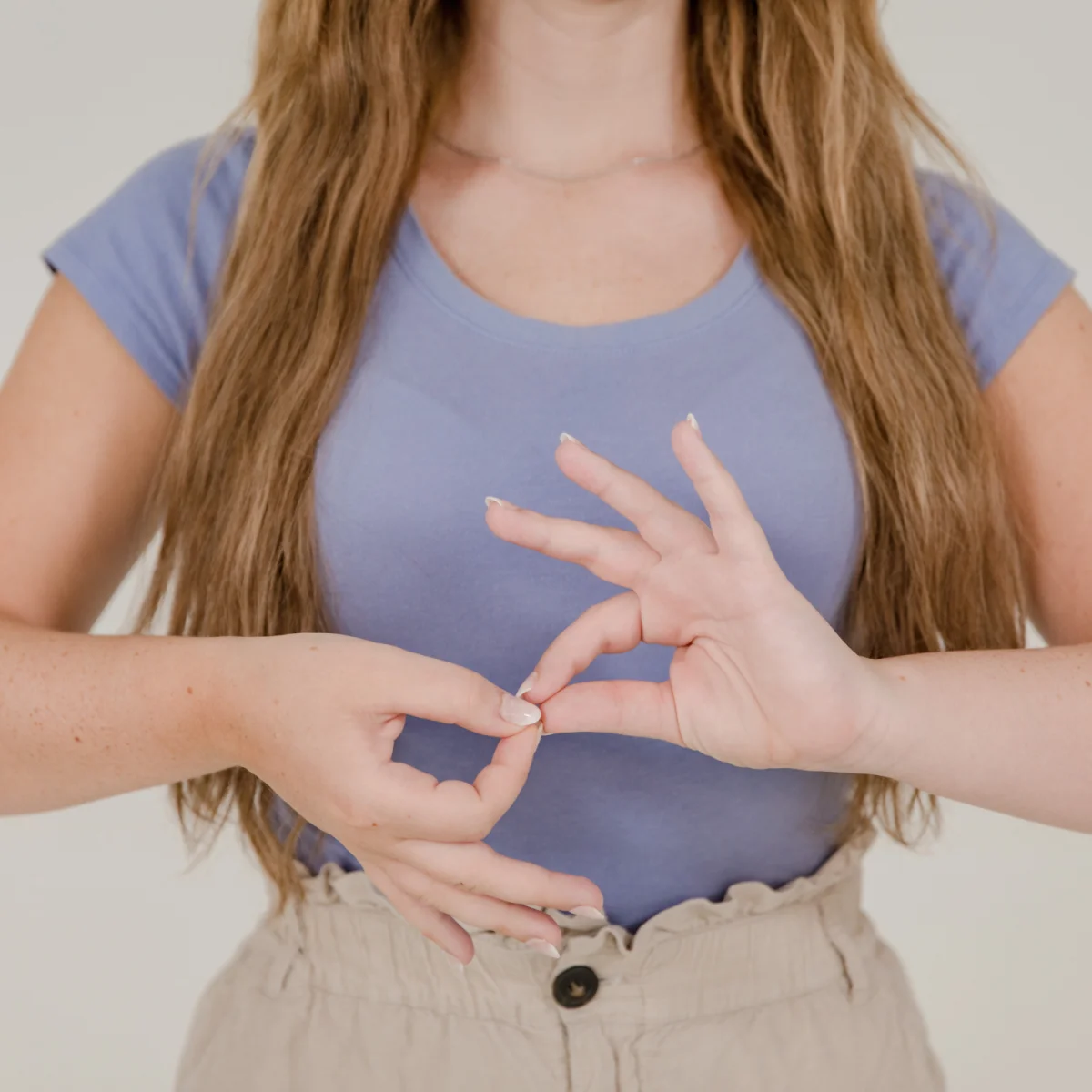 Woman demonstrating a hand gesture in sign language while standing against a neutral background.