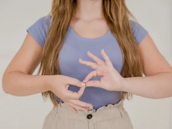 Woman demonstrating a hand gesture in sign language while standing against a neutral background.