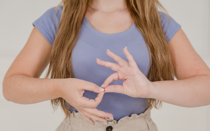 Woman demonstrating a hand gesture in sign language while standing against a neutral background.