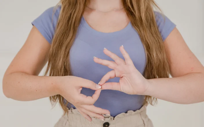 Woman demonstrating a hand gesture in sign language while standing against a neutral background.