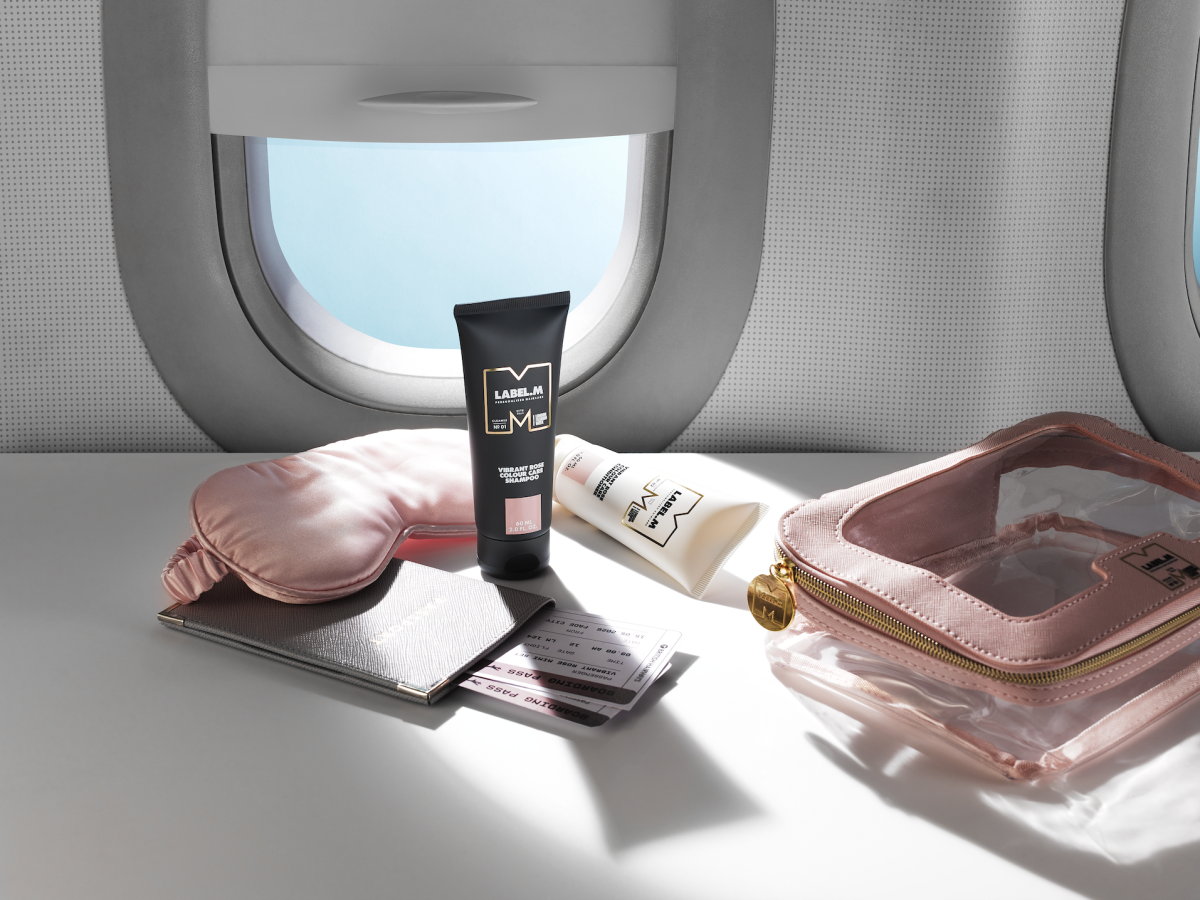LABEL.M travel haircare products displayed with a passport, sleep mask and travel pouch on an airplane tray table.