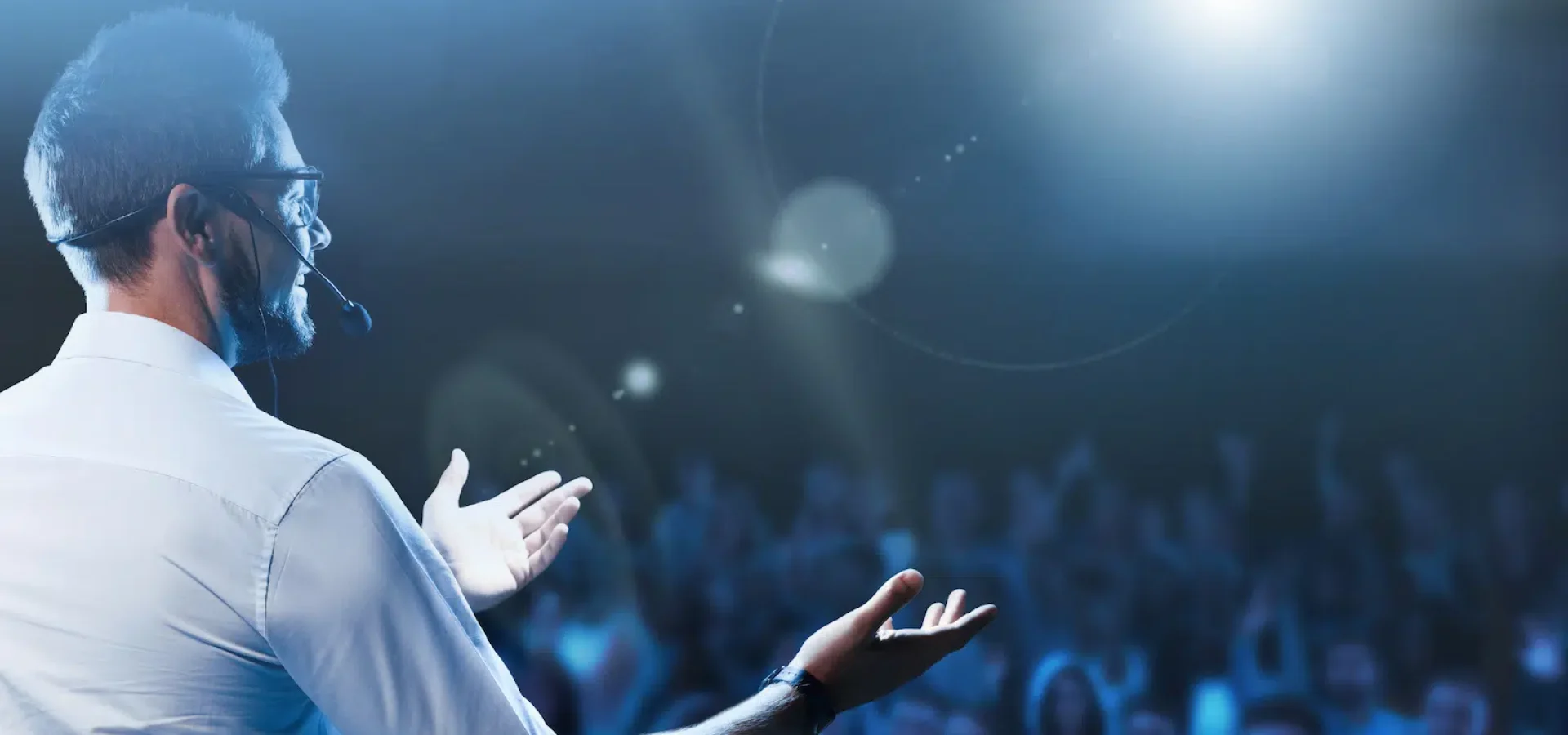 Conference speaker presenting on stage to a large audience with headset microphone and blue event lighting.