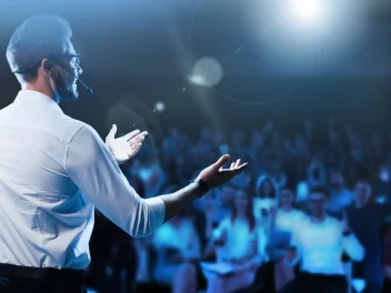 Conference speaker presenting on stage to a large audience with headset microphone and blue event lighting.
