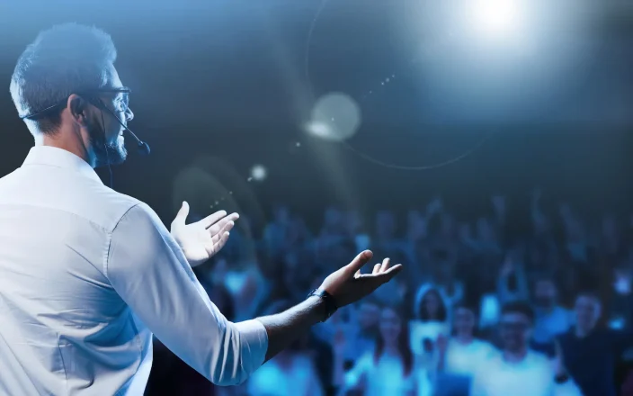 Conference speaker presenting on stage to a large audience with headset microphone and blue event lighting.