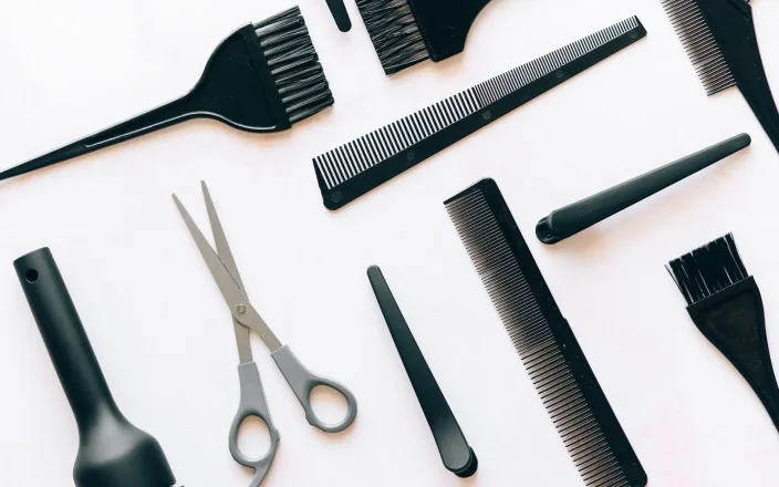 Flat lay of professional hairdressing tools including combs, tint brushes, scissors and styling equipment on white background.