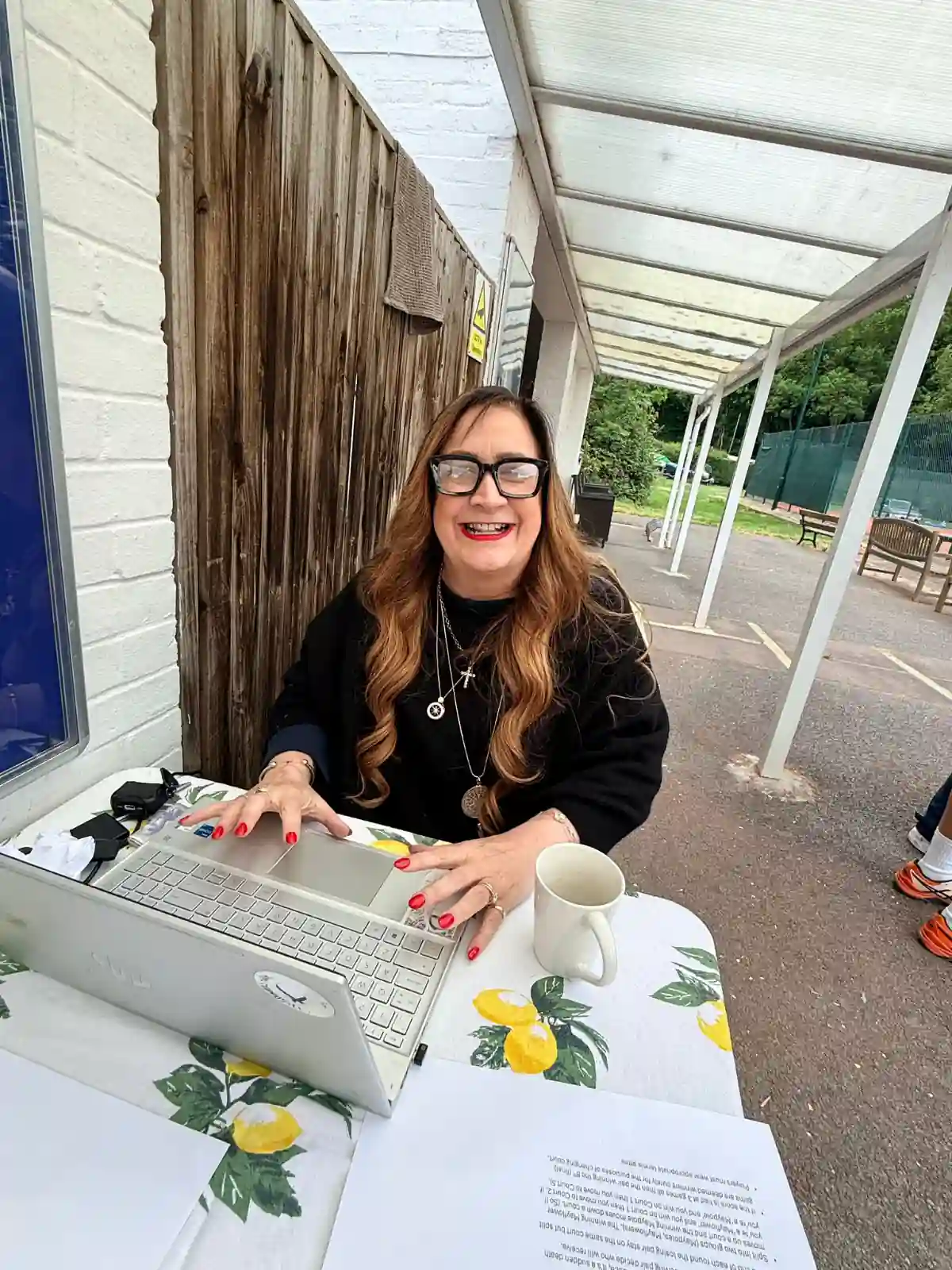 Woman with long wavy hair and glasses working on a laptop courtside at a Hair & Beauty Charity tennis event.