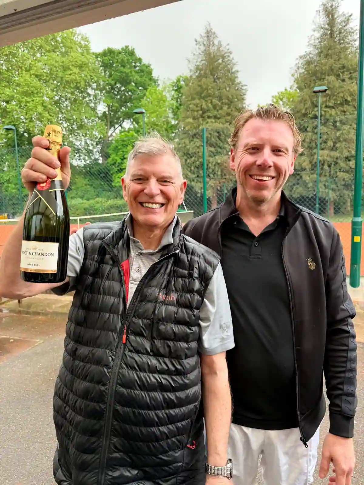 Two men smiling courtside at a Hair & Beauty Charity tennis event, holding a Champagne prize after the tournament.
