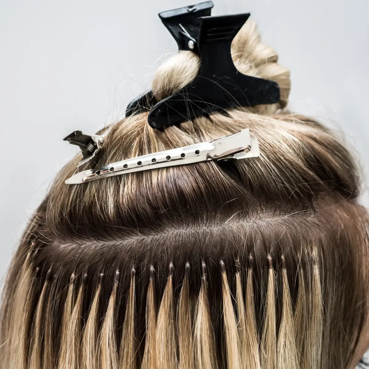 Close-up of blonde hair extensions being applied with sectioning clips and micro bonds during a salon fitting.
