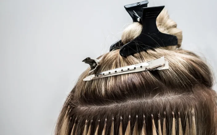 Close-up of blonde hair extensions being applied with sectioning clips and micro bonds during a salon fitting.