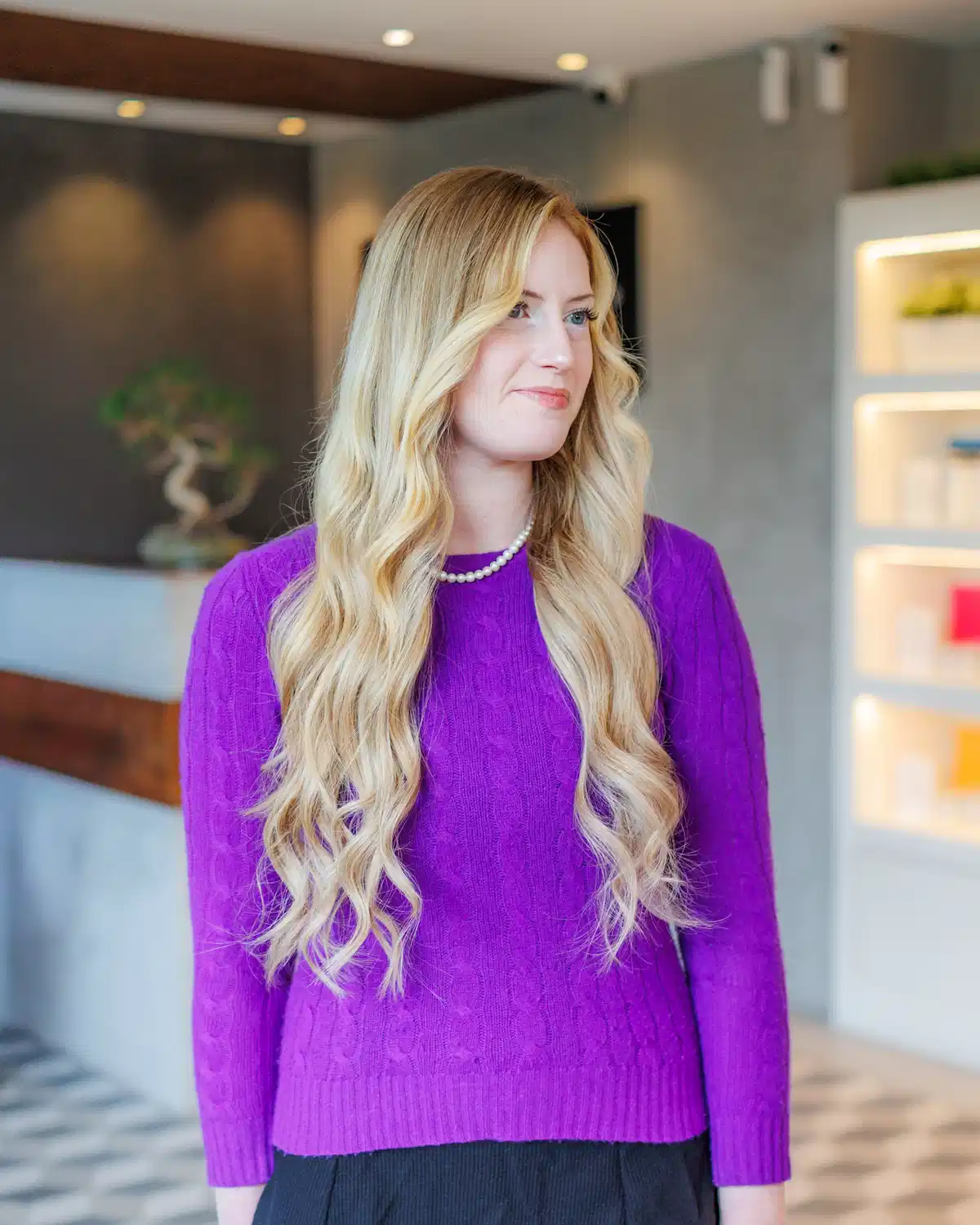 Woman with long blonde waves wearing a purple sweater, standing inside a modern hair salon.