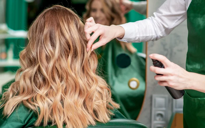 Stylist creating soft blonde waves with finishing spray during a salon hair styling appointment.