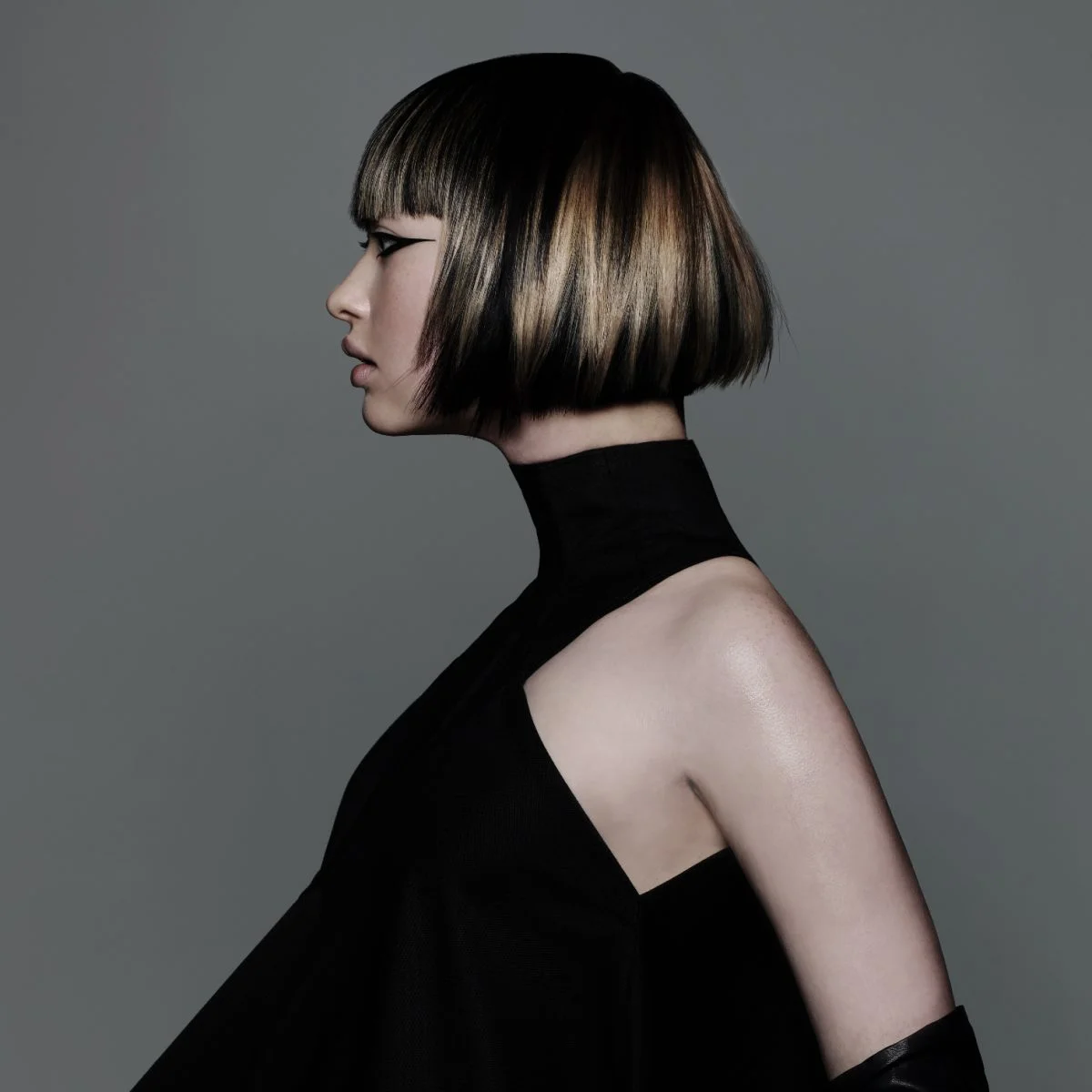 Model with sharp graphic bob haircut featuring blonde panel highlights and sleek precision styling against grey backdrop.