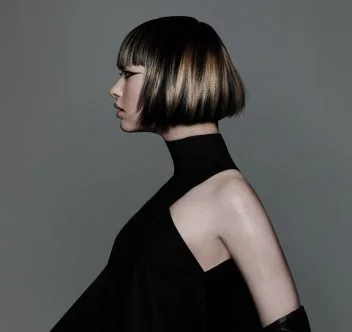 Model with sharp graphic bob haircut featuring blonde panel highlights and sleek precision styling against grey backdrop.
