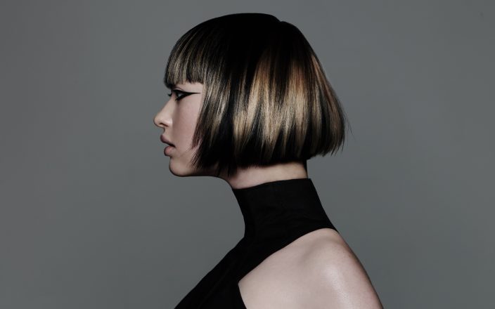 Model with sharp graphic bob haircut featuring blonde panel highlights and sleek precision styling against grey backdrop.