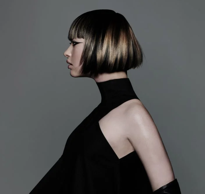 Model with sharp graphic bob haircut featuring blonde panel highlights and sleek precision styling against grey backdrop.