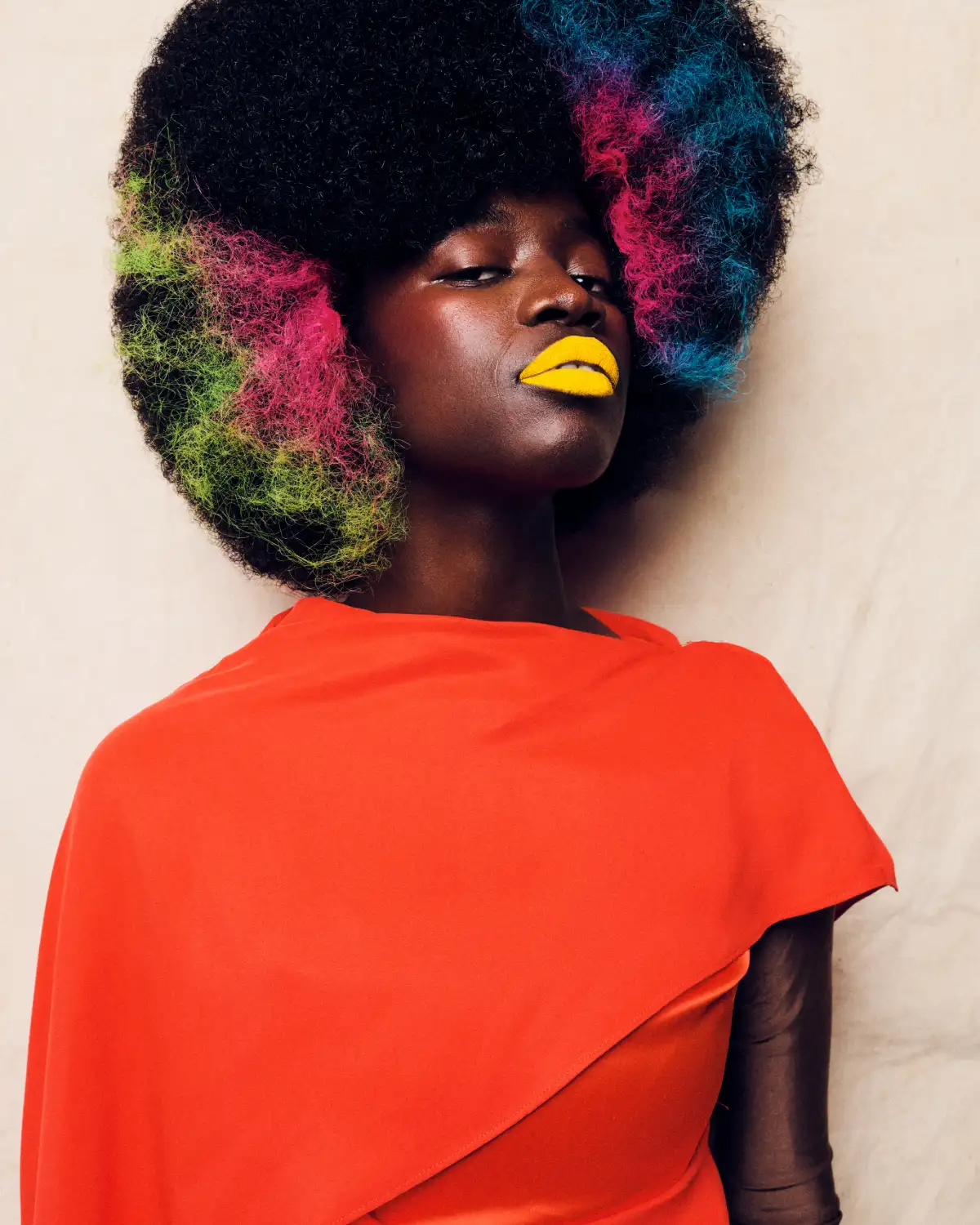 Model with a sculpted afro featuring vibrant pink, blue and green colour accents, paired with bold yellow lips in an editorial hair collection.