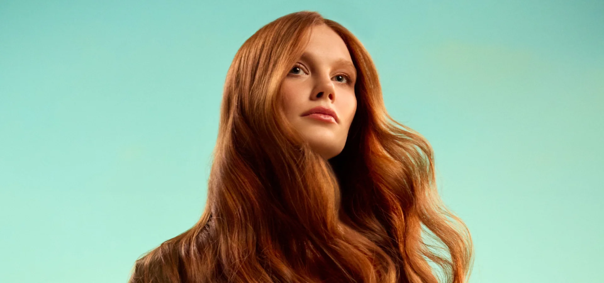 Model with long red hair stands in front of a green background as part of the JOICO Glow Warm campaign.