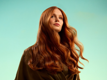 Model with long red hair stands in front of a green background as part of the JOICO Glow Warm campaign.