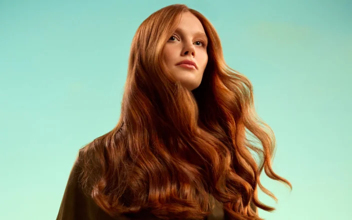 Model with long red hair stands in front of a green background as part of the JOICO Glow Warm campaign.