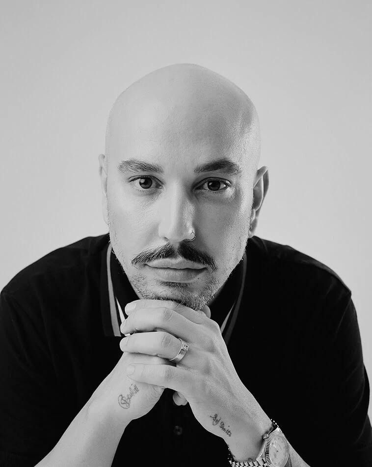 Portrait of hairstylist Pete Burkill in a black shirt, resting his chin on clasped hands in a black and white headshot.