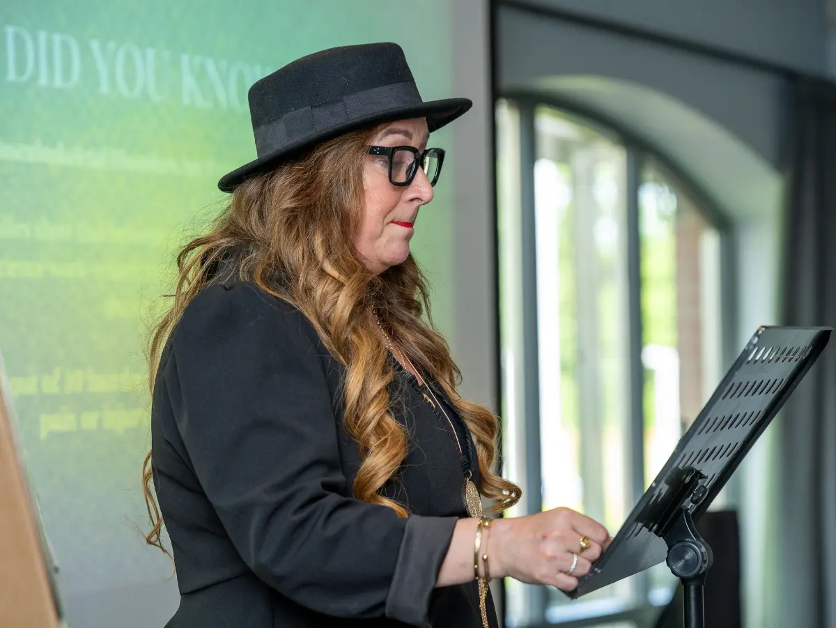Woman with long flowing brown hair with a top hat and glasses gives a speech at Never Just Hair exhibition.