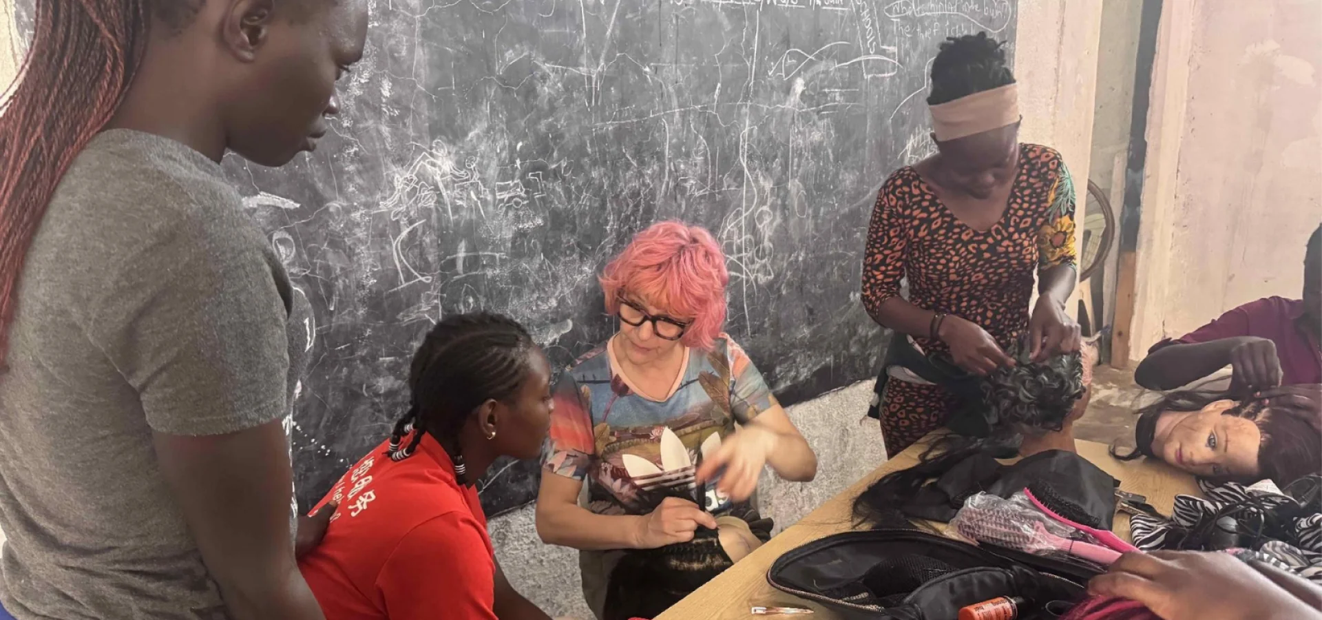 Anne Veck sits in front of a chalk board In Uganda teaching students hair skills.