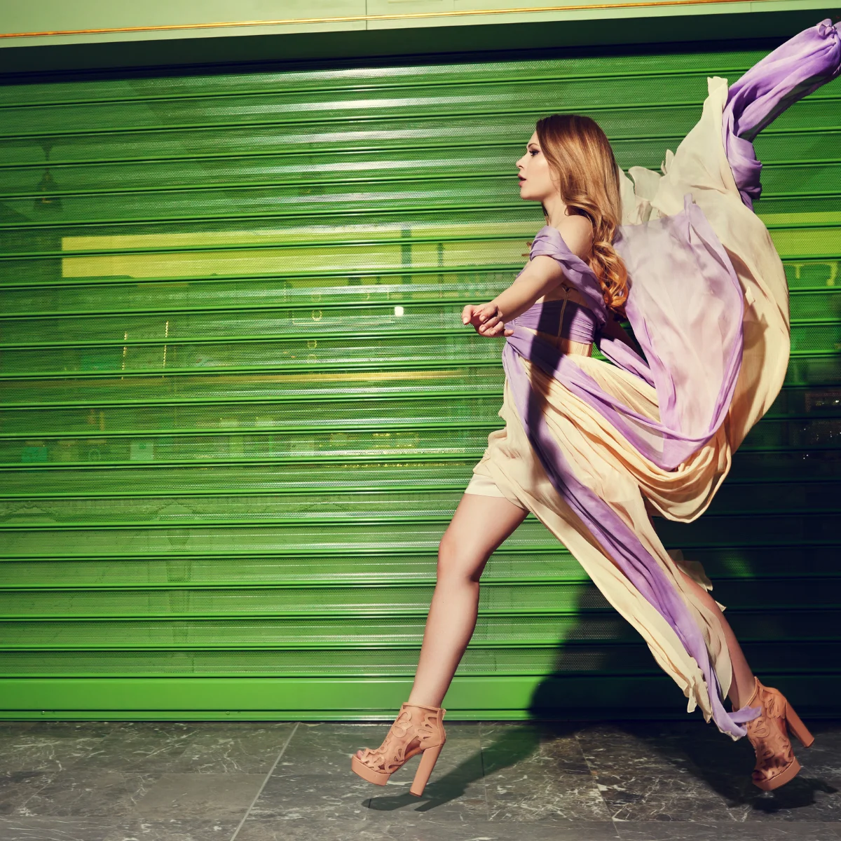 Model with long wavy brown hair walks pasta green garage door in a long dress being blown by the wind.