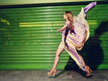Model with long wavy brown hair walks pasta green garage door in a long dress being blown by the wind.