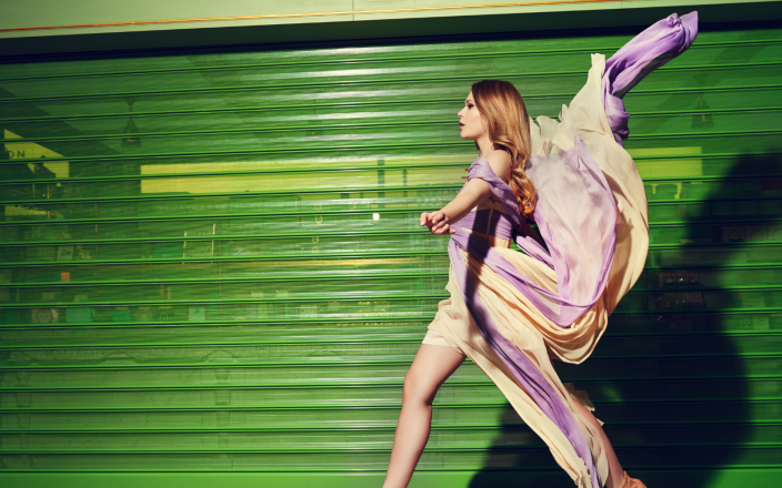 Model with long wavy brown hair walks pasta green garage door in a long dress being blown by the wind.