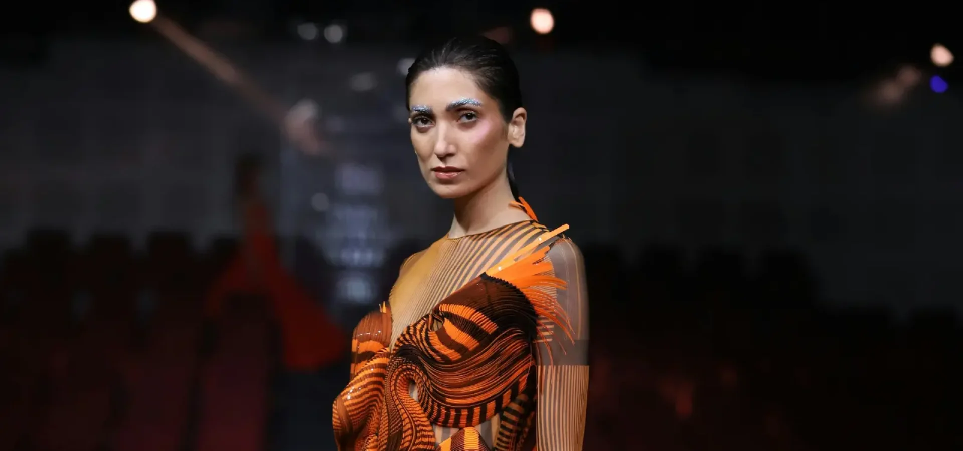 Model with slicked back hair and blue eyebrows on stage highlighting the F.A.M.E. Team finals.