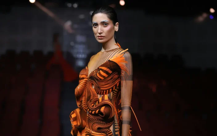 Model with slicked back hair and blue eyebrows on stage highlighting the F.A.M.E. Team finals.