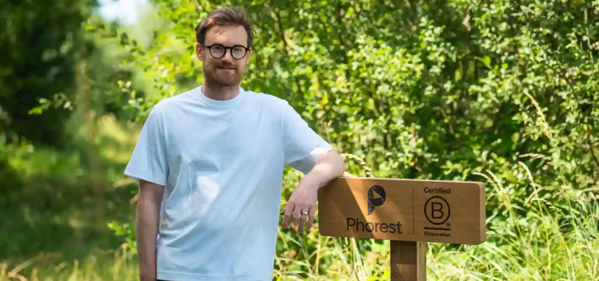 Ronan Perceval, Founder and CEO of Phorest B Corp stands in a blue t-shirt next to sign in the forest.