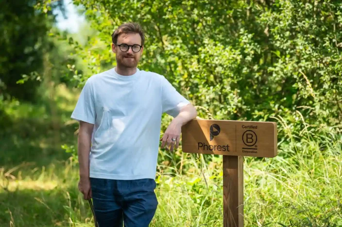 Ronan Perceval, Founder and CEO of Phorest B Corp stands in a blue t-shirt next to sign in the forest.