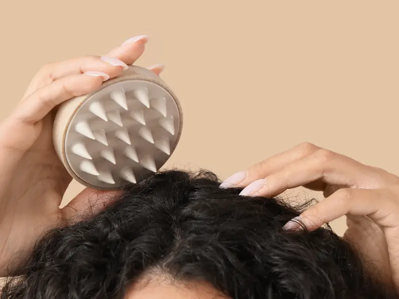 Hands using a hand-held scalp massager tool with soft silicone designed to boost blood flow, exfoliate product buildup, and help lather shampoo during winter.
